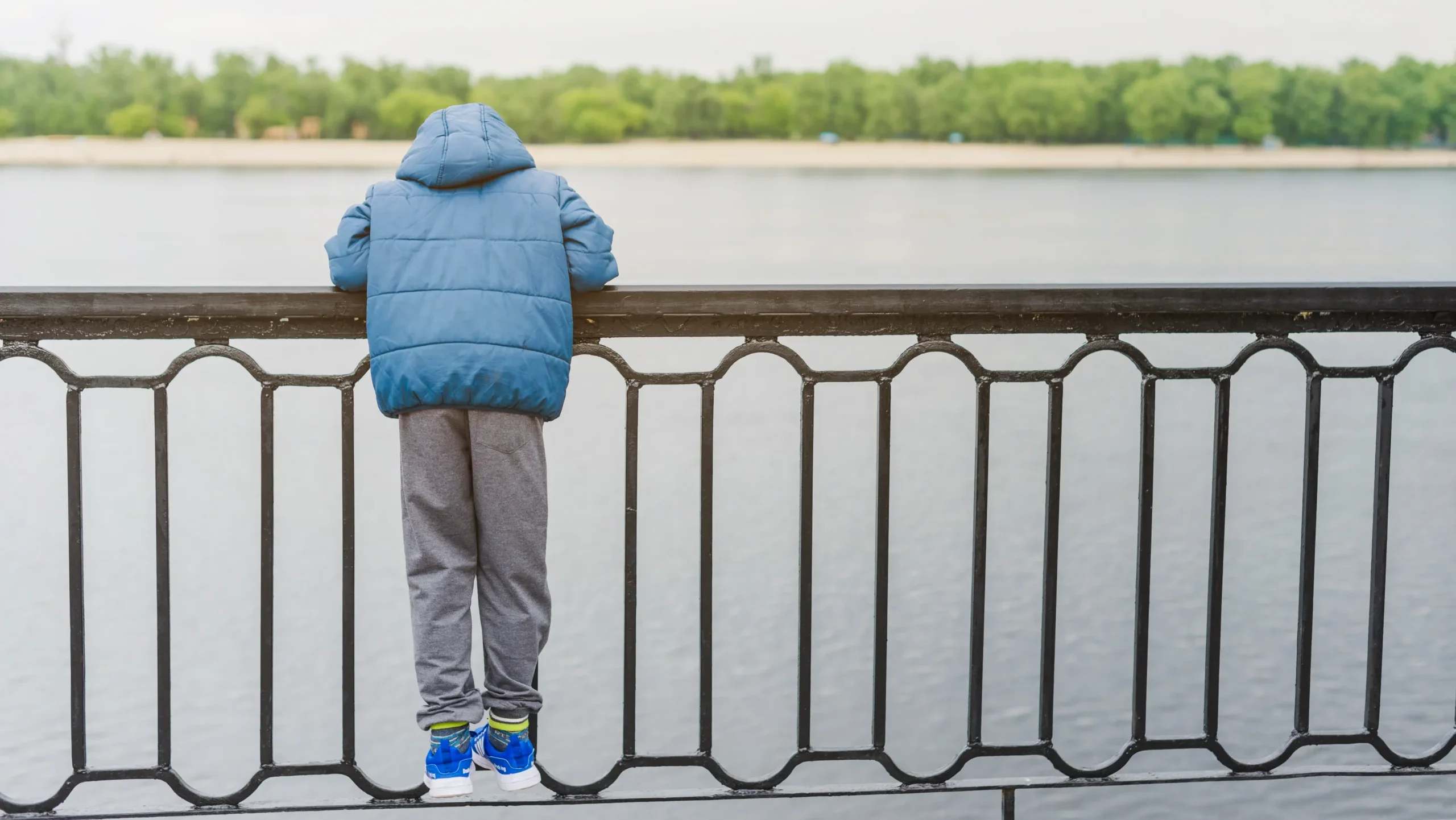 Boy in blue jacket leaning on black metal fence overlooking water, best fence material